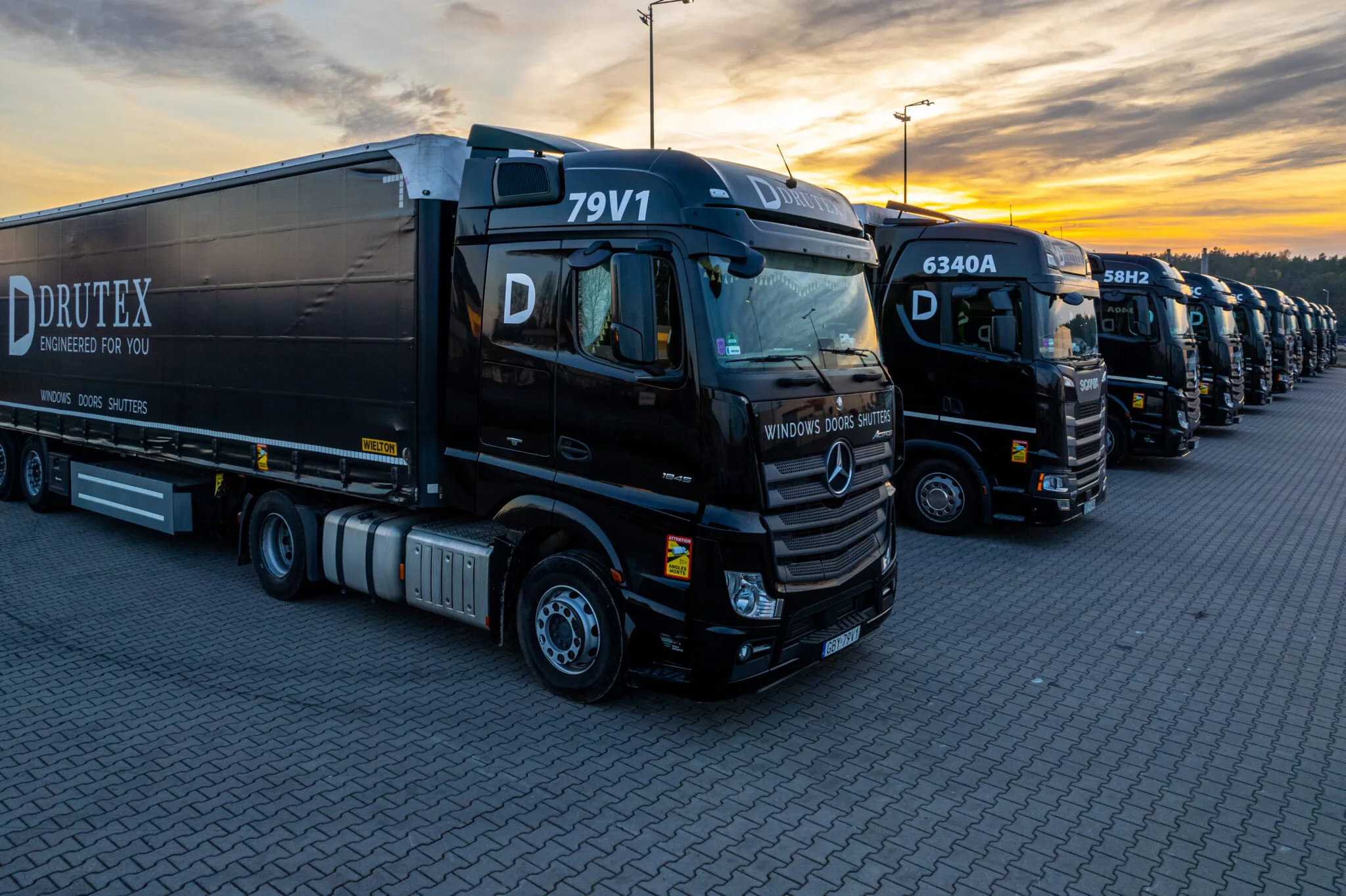 Eine Reihe von schwarzen Lastwagen der Firma DRUTEX, aufgereiht auf einem Parkplatz bei Sonnenuntergang