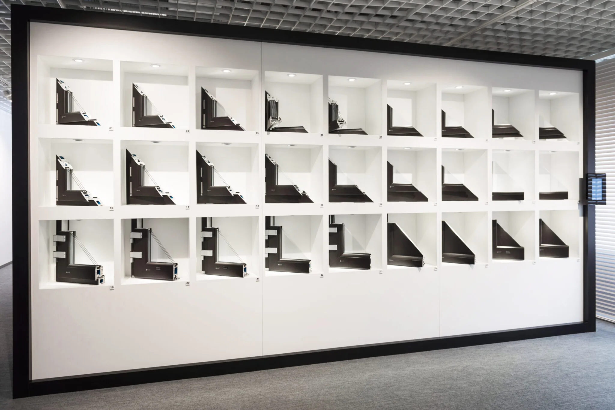 Ein Ausstellungsraum mit verschiedenen Fenstermodellen in weißen Rahmen, die auf einer großen Wand montiert sind